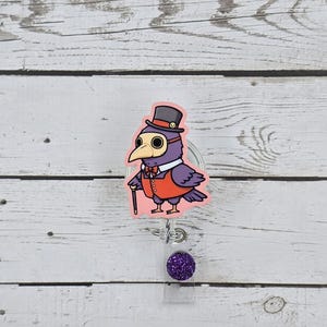 May include: A purple and black cartoon bird wearing a top hat, goggles, a bow tie, and a red vest. The bird is holding a cane and is standing on a pink background. The bird is attached to a clear plastic retractable badge holder with a purple glitter button.