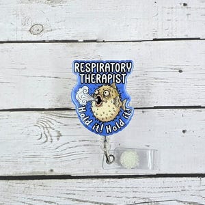 May include: A blue badge reel with the text "RESPIRATORY THERAPIST" and a cartoon pufferfish blowing out air. The badge also says "Hold it!" on either side of the fish. The reel has a clear plastic holder and a silver clip.