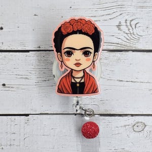 May include: A pink and clear plastic retractable badge holder with a cartoon image of Frida Kahlo wearing a red shawl and a flower crown. The badge holder has a red glitter ball on the end of the retractable cord.