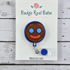 May include: A brown and blue lollipop badge reel with a smiling face. The reel is attached to a blue glitter charm. The card reads "Badge Reel Babe" and "Handmade with Love".