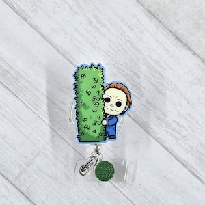 May include: A retractable badge reel featuring a cartoon illustration of a character peeking from behind a green bush. The character is wearing a blue jumpsuit. The reel has a clear plastic base with a green glitter accent.