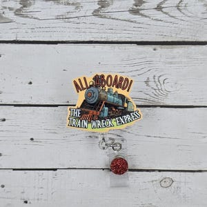 May include: A yellow and blue badge reel with a cartoon train and the text "All Aboard! The Train Wreck Express". The reel has a red glitter charm.