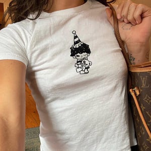 May include: White short-sleeved t-shirt featuring a black and white cartoon graphic of a character wearing a party hat and holding a teddy bear. The shirt is a fitted style.