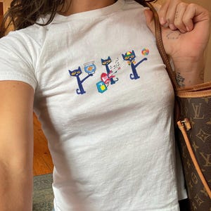 May include: White crew neck t-shirt with a colorful graphic of three stylized blue cats. The cats are holding a fishbowl, a gift, and a lollipop. The word "PETY" is printed above the cats in a matching font.