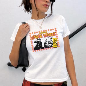 May include: A white short-sleeved t-shirt featuring a graphic print with the text CHUCK TIMELY and THE THIRD ALBUM. The design includes a yellow star border, a black and white illustration of two figures, and a yellow hourglass icon on the right.
