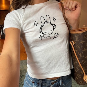 May include: White t-shirt featuring a black graphic of a bunny wearing headphones and stars. The top is paired with blue jeans. A brown handbag with a monogram pattern is visible.