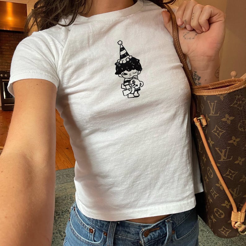 May include: White short-sleeved t-shirt featuring a black and white cartoon graphic of a character wearing a party hat and holding a teddy bear. The shirt is a fitted style.