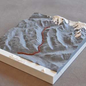 Puede incluir: Un modelo 3D gris de una cordillera con un sendero rojo que serpentea a través de ella. El modelo está sobre una base blanca.