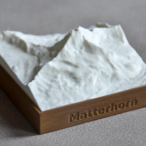 以下が含まれることがあります： マッターホルンの山頂を再現した白い3Dプリントの地形モデルで、詳細な地形の質感が特徴です。このモデルは正方形の茶色の台座に取り付けられており、木製フレームの前面にはMatterhornという文字が刻印されています。