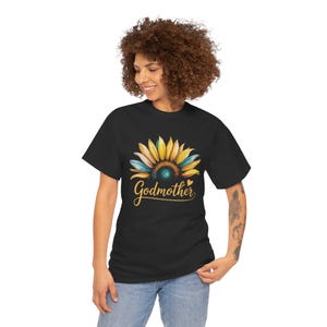 Puede incluir: Camiseta negra con un diseño de girasol en acuarela con pétalos amarillos, azules y turquesas. La palabra "Godmother" está escrita en cursiva debajo de la flor. La camiseta es de estilo casual.