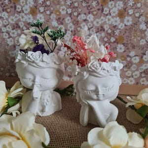 Peut inclure: Deux pots de fleurs en céramique blanche en forme de visage féminin aux yeux fermés. Chaque pot est surmonté d'une couronne de fleurs et rempli de fleurs séchées. Les fleurs du pot de gauche sont violettes, blanches et vertes. Les fleurs du pot de droite sont roses et blanches.