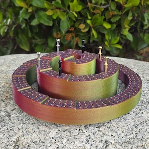May include: A spiral cribbage board with a gradient of colors from red to green. The board has multiple levels and is dotted with small holes. Several metal pegs are inserted into the holes.