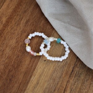 Könnte beinhalten: Zwei weiße Perlenringe mit kleinen bunten Edelsteineinsätzen. Die Ringe liegen auf einer Holzoberfläche.