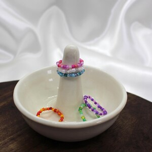 Könnte beinhalten: Ein weißer Keramik-Ringhalter mit einem zentralen Pfosten, der mit Perlen besetzte Ringe in Rosa, Blau und Weiß hält. Zusätzliche Perlenringe in Orange, Grün und Lila liegen in der Schale. Der Hintergrund ist ein weißer, seidiger Stoff.
