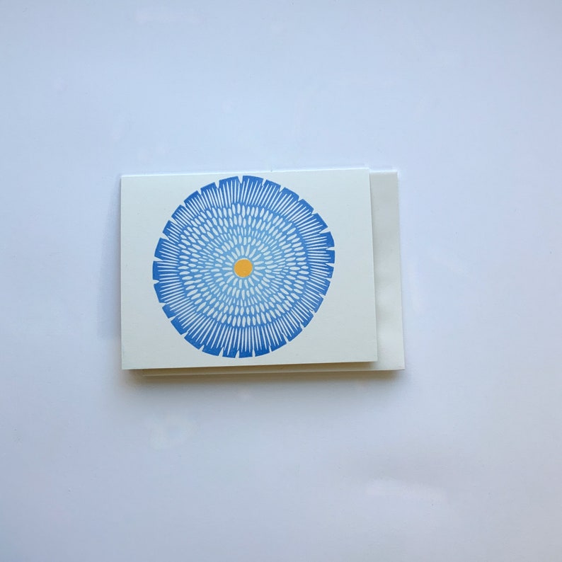 Op de afbeelding: Een witte kaart met een blauw en wit cirkelvormig ontwerp met een geel centrum. Het ontwerp lijkt op een bloem.