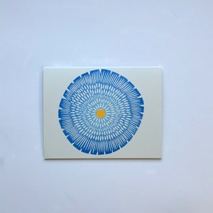 Op de afbeelding: Een blauw en wit cirkelvormig ontwerp met een geel centrum. Het ontwerp doet denken aan een bloem of een mandala.