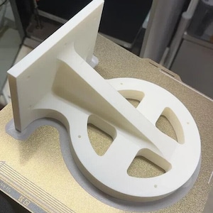 Pode incluir: Um objeto branco impresso em 3D com uma base circular e uma seção elevada em ângulo. O objeto tem vários furos na base e na seção em ângulo.