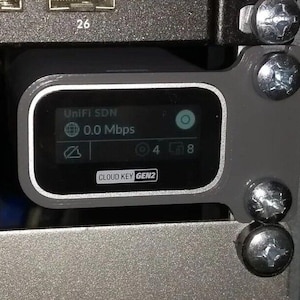 May include: A gray UniFi SDN Cloud Key Gen2 device mounted on a wall with a black screen displaying "UniFi SDN" and "0.0 Mbps".