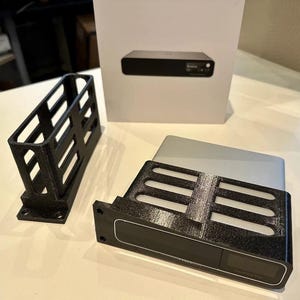 Op de afbeelding: Twee zwarte 3D-geprinte standaards voor een klein elektronisch apparaat. De standaard aan de linkerkant is een verticale standaard met een rechthoekige basis. De standaard aan de rechterkant is een horizontale standaard met een rechthoekige basis en een verhoogde lip aan de voorkant. De standaard aan de rechterkant heeft een klein zwart scherm met de tekst "Sound BlasterX Kratos S5" erop.