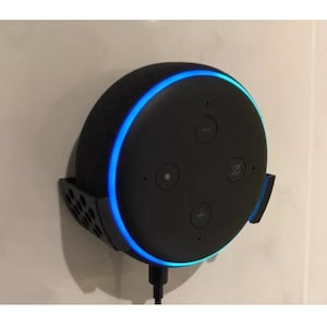 Peut inclure: Un haut-parleur intelligent noir avec un anneau lumineux bleu autour du bord. Le haut-parleur est monté sur un mur avec un support noir.