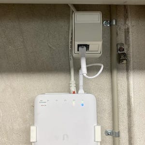 以下が含まれることがあります： 青と緑のライトインジケーターが付いた白いルーター。ルーターは白いブラケットで壁に取り付けられています。白いケーブルがルーターに接続されています。