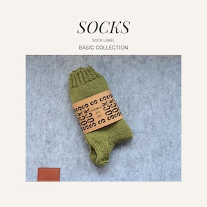 Op de afbeelding: Een olijfgroene sok met een bruine papieren band eromheen. Op de band staat het woord 'SOCKS' meerdere keren afgedrukt. De sok maakt deel uit van de 'Basic Collection' en wordt gepresenteerd op een grijze ondergrond.