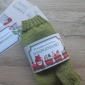 Puede incluir: Un par de calcetines de punto verde oliva con una banda decorativa con un tren navideño con gnomos. La banda dice "HANDGEMAAKT". Se ven dos tarjetas con instrucciones de lavado.