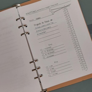 Op de afbeelding: Open plannerpagina met de titel "KNITTING: MONTHLY HABIT TRACKER". De pagina bevat een lijst met breiprojecten en een sleutel voor tijd en rijen. Een breisteekgrafiek loopt aan de rechterkant.