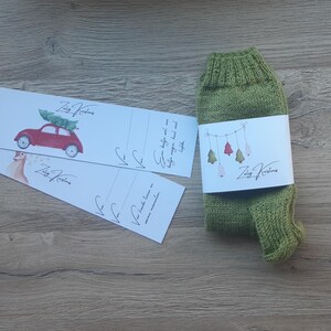 Puede incluir: Un par de calcetines de punto verde oliva con una etiqueta blanca con ilustraciones de árboles de Navidad. La etiqueta también incluye el texto "Zalig Kerstmis". También hay tres tarjetas blancas con ilustraciones.
