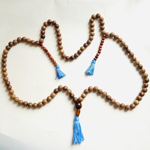 Puede incluir: Un collar de cuentas de madera con borlas azules. Las cuentas son marrones y tienen una veta de madera natural. El collar mide aproximadamente 100 cm de largo.