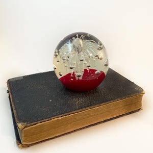 Pode incluir: Um peso de papel de vidro transparente com uma base vermelha e um design branco e pontiagudo no interior. O peso de papel está em cima de um livro antigo, encadernado em couro preto, com páginas amareladas. O peso de papel tem bolhas de ar.