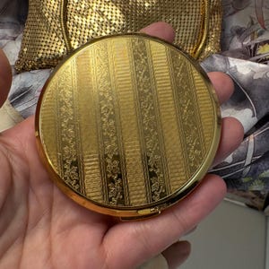 May include: A round gold-tone metal compact mirror featuring an engraved surface with alternating vertical stripes of textured cross-hatching and delicate floral vine patterns. The compact has a polished finish and a hinged closure mechanism.