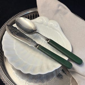 May include: A silver spoon and butter knife with dark green handles rest on a white, scalloped dish. The dish sits on a silver serving tray, with a linen napkin in the background. The spoon has a decorative floral design.