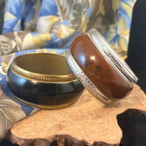 Puede incluir: Dos pulseras gruesas. Una es negra con ribete dorado, la otra marrón con ribete plateado. Las pulseras se exhiben sobre un trozo de madera, con una tela estampada en el fondo.