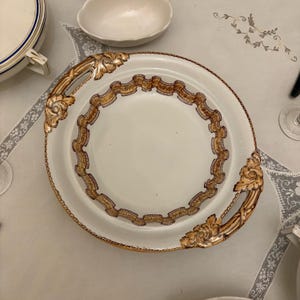 Puede incluir: Una fuente blanca con asas doradas y un borde decorativo dorado y marrón. La fuente tiene un diseño central y forma parte de un juego de vajilla. Las asas tienen detalles florales ornamentados.