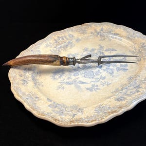 May include: An antique carving fork with a brown antler handle and a silver-toned metal fork. The fork rests on a vintage oval serving platter with a blue floral pattern. The platter has a cream-colored background.