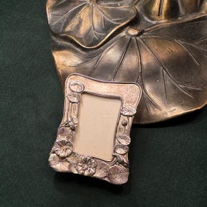 May include: A rectangular silver-toned metal picture frame featuring a relief design of lily pads and lotus flowers along the border. The frame has a smooth, light-colored center insert and is positioned in front of a large, dark bronze-colored metal lotus leaf.