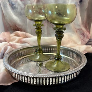 May include: Two olive green glass goblets with rounded bowls and twisted stems, positioned on a round silver-toned metal tray featuring a decorative pierced gallery rim and an intricate engraved pattern on the flat interior surface.