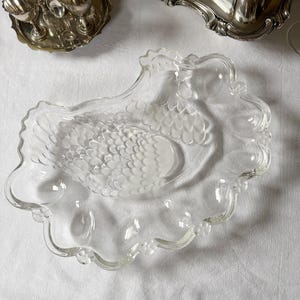 Peut inclure: Plat de service en verre transparent avec un motif de cygne. Le plat a un bord festonné et une image de cygne givré au centre. D'autres pièces de service en argent sont visibles en arrière-plan.