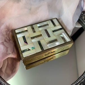 May include: A rectangular, vintage jewelry box with a brass frame and a mosaic top. The mosaic is made of rectangular pieces of iridescent mother-of-pearl, arranged in a woven pattern. The box is closed.
