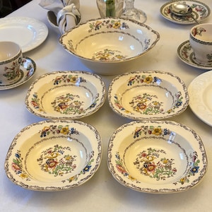 May include: A collection of vintage cream-colored china with floral patterns. The set includes a large serving bowl, several smaller bowls, teacups, saucers, and plates. The floral design features yellow, pink, and blue flowers with brown trim. A classic tableware set.