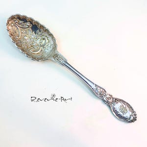 Puede incluir: Una cuchara de servir de plata con un diseño floral ornamentado. La cuchara tiene un cuenco redondeado y un mango largo y delgado.
