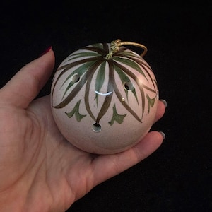 May include: A round, ceramic musical instrument with a light pink base color. The instrument features a decorative pattern of green and brown leaf-like designs. It has several holes and a gold-colored cord attached at the top.