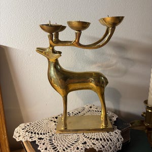 May include: A polished brass candelabra shaped like a standing deer. The deer features an elongated neck and head, with its antlers extending upwards to form three distinct candle holders. The entire structure is mounted on a rectangular base.