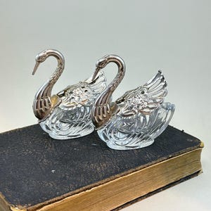 May include: Two vintage swan-shaped salt and pepper shakers. The shakers have clear glass bodies with silver-colored metal swan heads and wings. The shakers are resting on a dark, textured surface, likely a book.