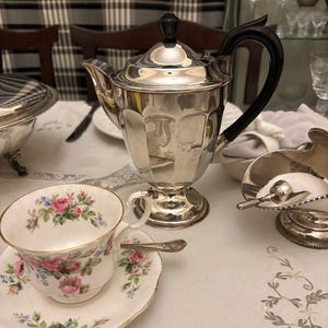 Può includere: Un servizio da tè in argento con manico e coperchio neri, una tazza e un piattino floreali e una zuccheriera con cucchiaio sono disposti su una tovaglia bianca. La tazza e il piattino presentano rose rosa e blu.