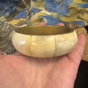 May include: Cream-colored bangle bracelet with a brass rim. The bracelet is made of rectangular segments. The background includes a floral patterned fabric.