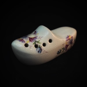 May include: A ceramic clog with a floral design in purple, blue, and yellow. The off-white shoe has three holes and a crackled glaze. The decorative Dutch shoe is set against a black background.