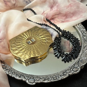 May include: A gold compact with the letters "H S" is displayed on a silver-rimmed mirror. A black beaded necklace and a black floral necklace are also on the mirror. A pink and white floral scarf is in the background.