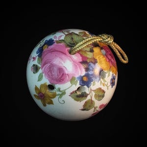 May include: A ceramic ocarina with a floral design featuring pink roses, yellow and blue flowers, and green leaves. The ocarina has a gold cord and is set against a black background.